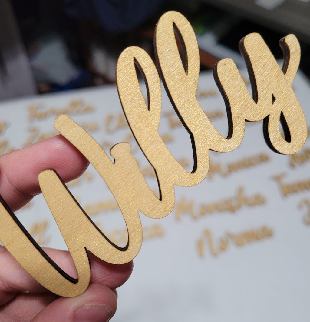 A hand holding a gold laser-cut wooden name sign that reads 'Willy' in a cursive script, with a blurred background showing other engraved names on a white surface.