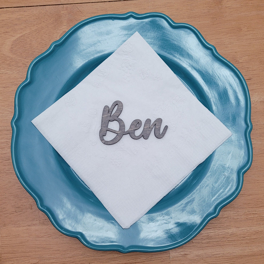 A white paper napkin with the name 'Ben' in metallic script, placed on a scalloped teal blue plate on a wooden table. Other metallic script name cutouts are visible at the bottom of the frame.