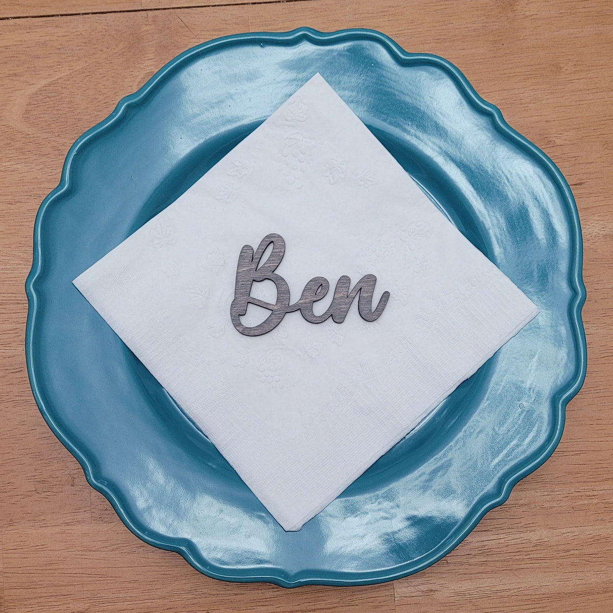 A white paper napkin with the name 'Ben' in metallic script, placed on a scalloped teal blue plate on a wooden table. Other metallic script name cutouts are visible at the bottom of the frame.