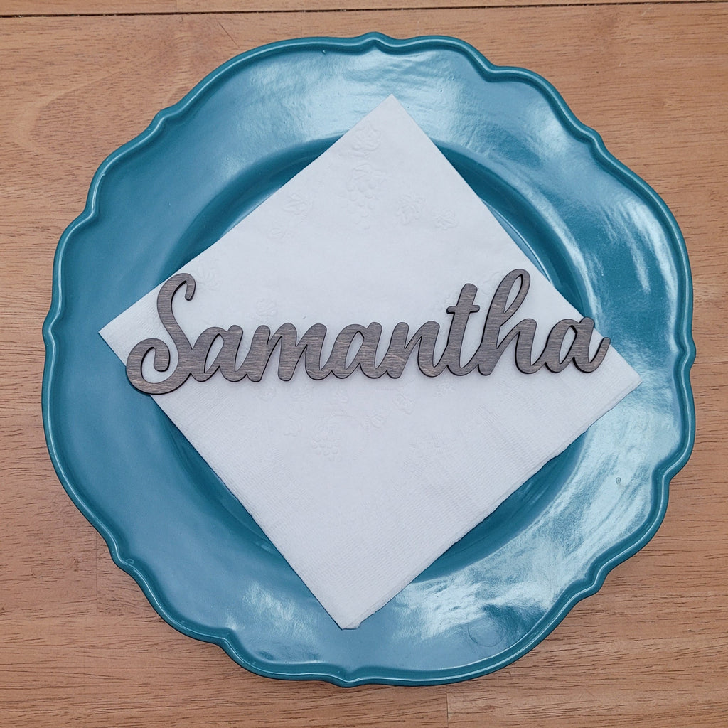 A personalized place setting featuring a scalloped teal blue plate, a white folded napkin, and a silver metal script nameplate reading 'Samantha', displayed on a wooden tabletop.