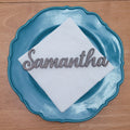 A personalized place setting featuring a scalloped teal blue plate, a white folded napkin, and a silver metal script nameplate reading 'Samantha', displayed on a wooden tabletop.