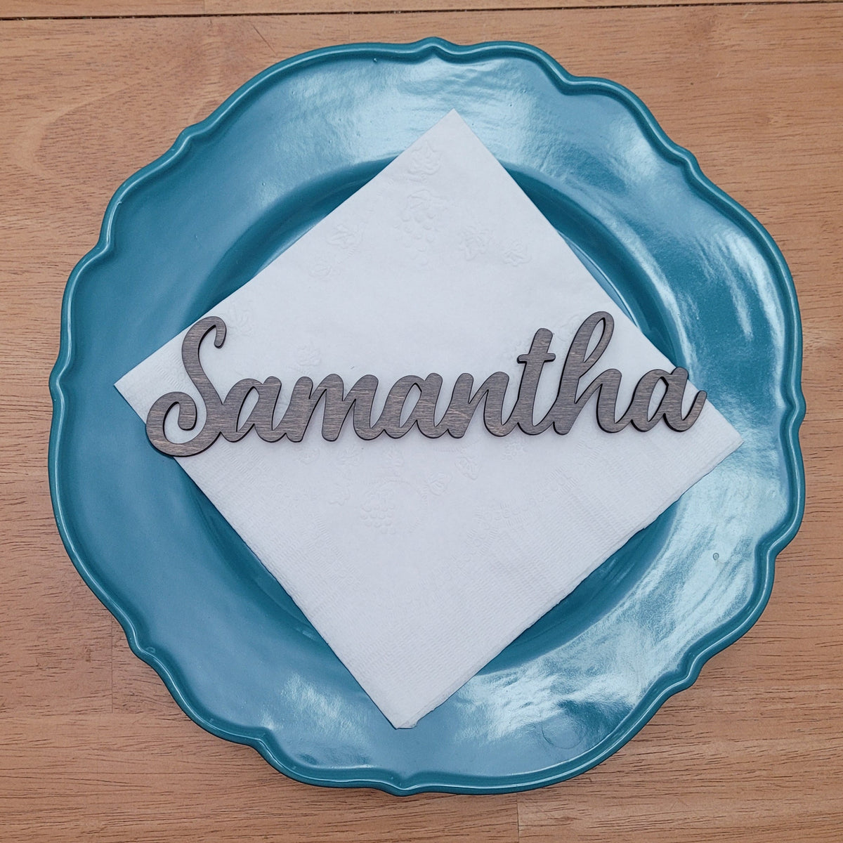 A personalized place setting featuring a scalloped teal blue plate, a white folded napkin, and a silver metal script nameplate reading 'Samantha', displayed on a wooden tabletop.