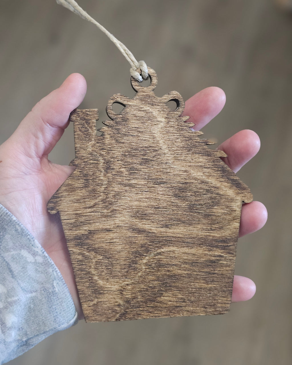 A hand holding a rustic, espresso finished wooden house-shaped ornament with a natural wood grain texture and a small hanging twine string.