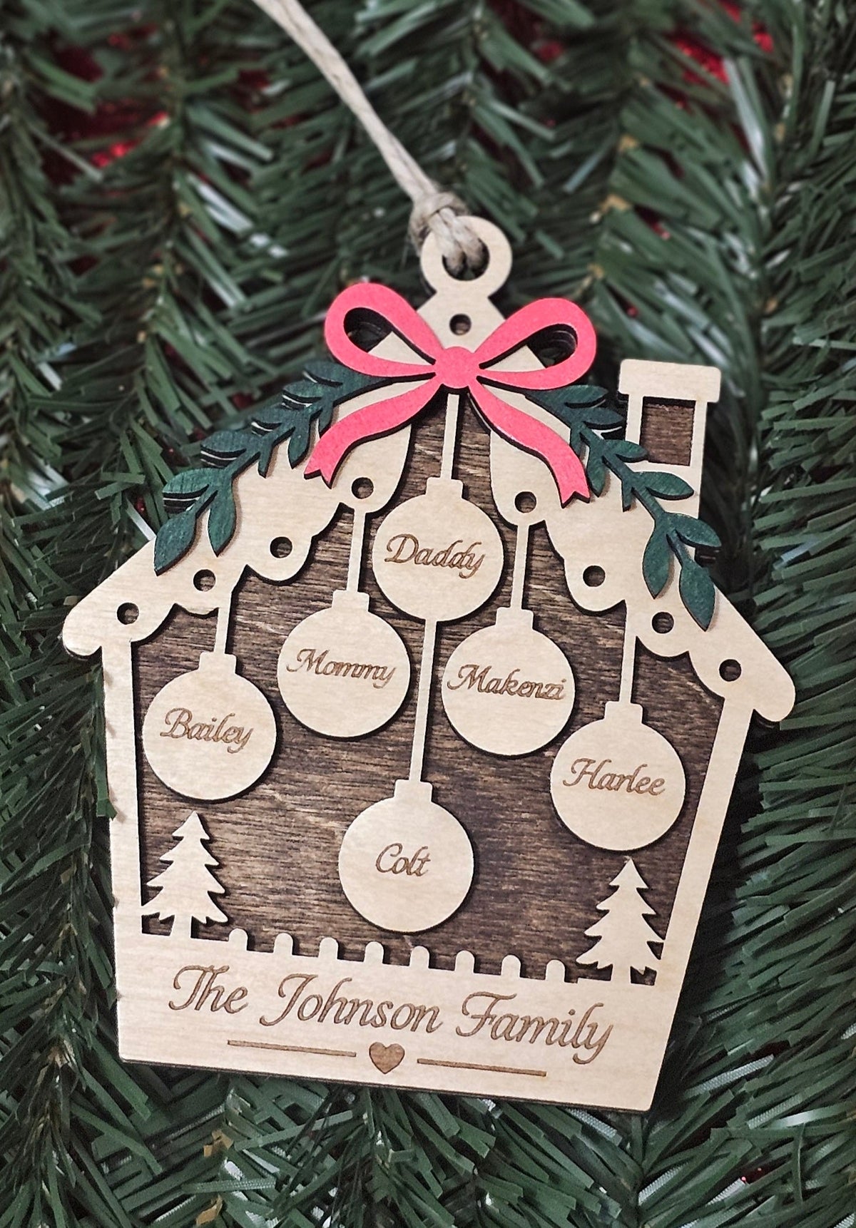 A personalized wooden house Christmas ornament hanging on a green Christmas tree. The ornament features a light natural wood body with a dark wood-grain printed center panel, a pink bow at the top, and laser-engraved names including Bailey, Mommy, Makensie, Harloe, Colt, and The Johnson Family.