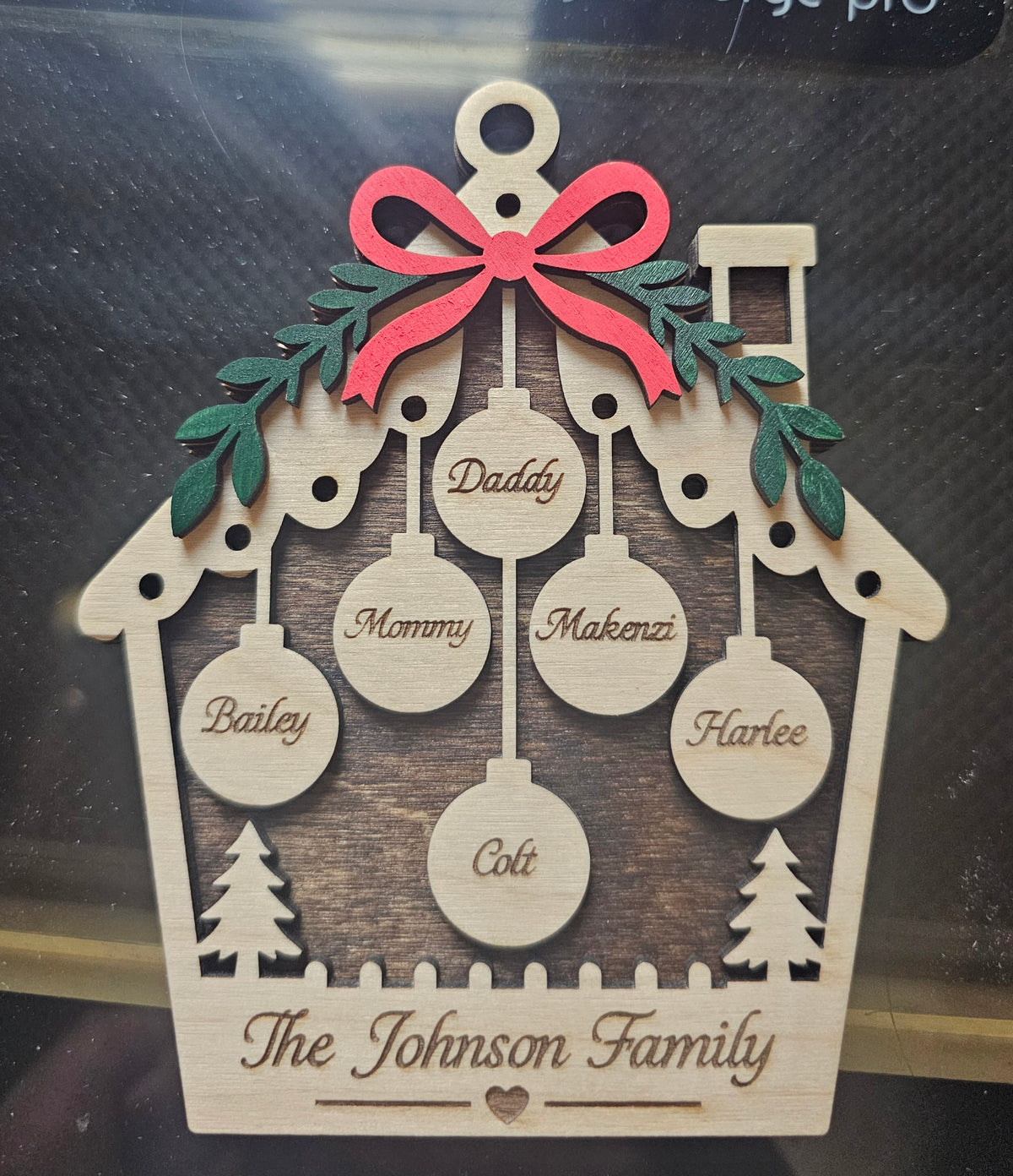 A personalized wooden Christmas ornament shaped like a house with a red bow, green holly leaves, and laser-engraved names of family members including Daddy, Mommy, Makenzi, Bailey, Harlee, Coli, and The Johnson Family.