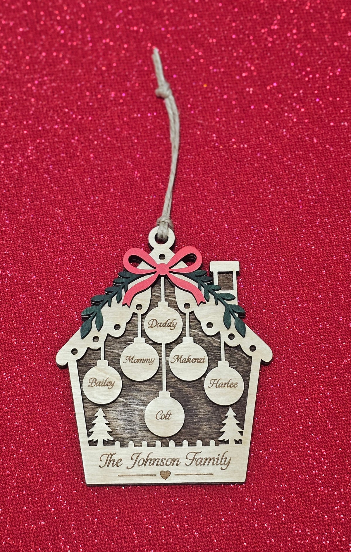 A personalized finished wooden house Christmas ornament with a twine hanging string, featuring laser-engraved family names inside hanging circles, a red bow, and green pine branches, displayed on a red glittery background.