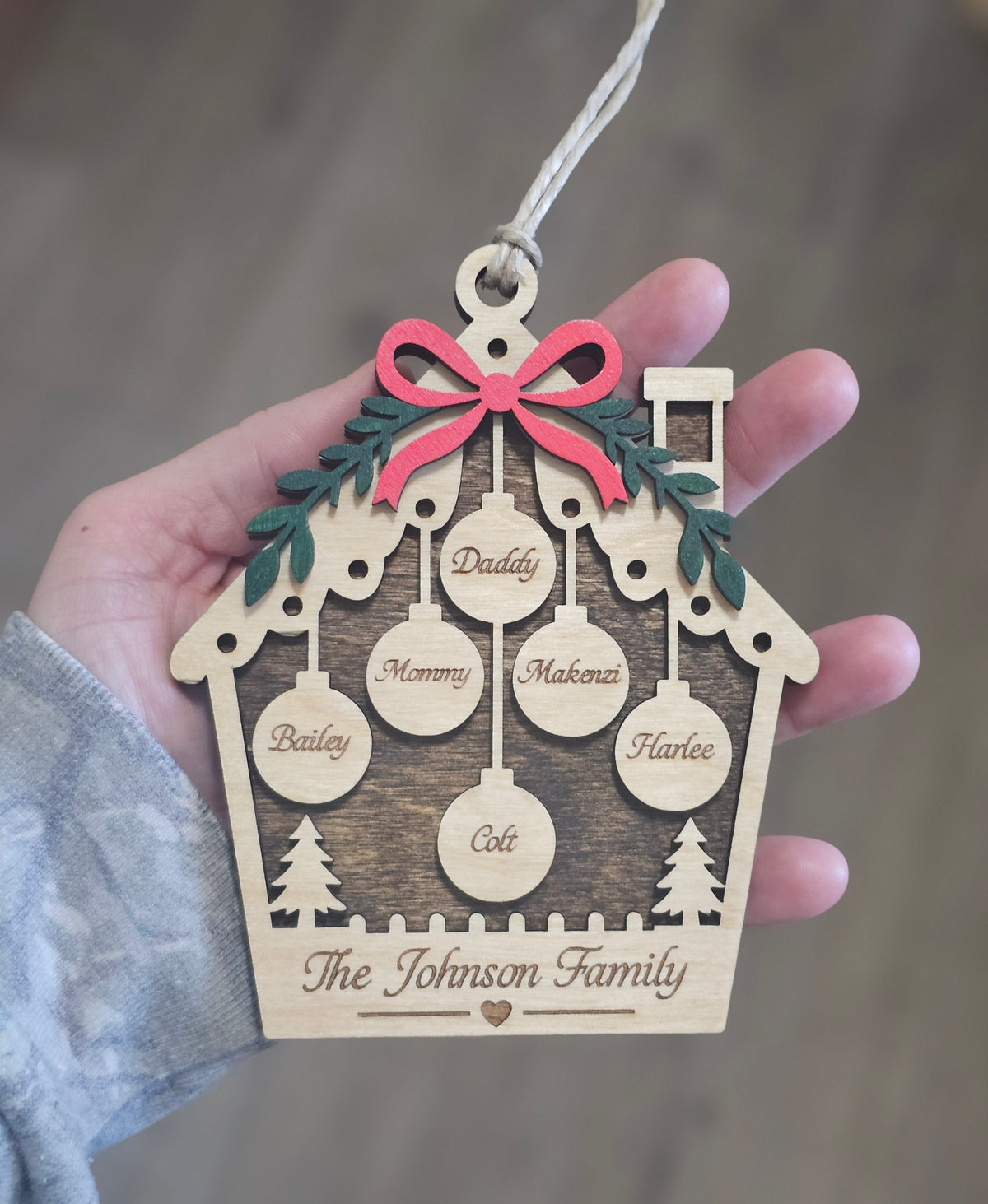 A hand holding a personalized wooden house Christmas ornament with laser-engraved names and a pink bow, shown against a blurred indoor background.
