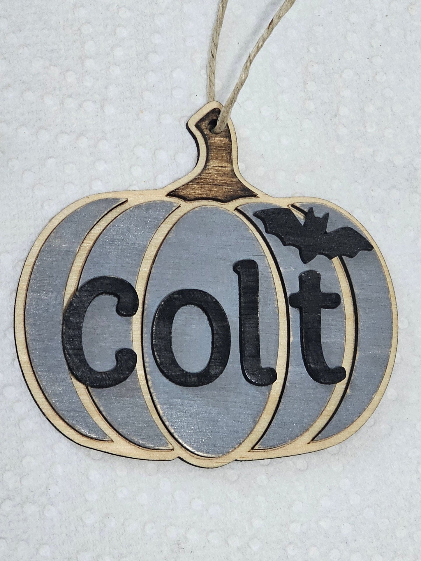 A wooden hanging pumpkin ornament with a gray-painted finish, black 'colt' lettering, a small black bat silhouette, and a natural wood stem, suspended by twine on a white textured background.