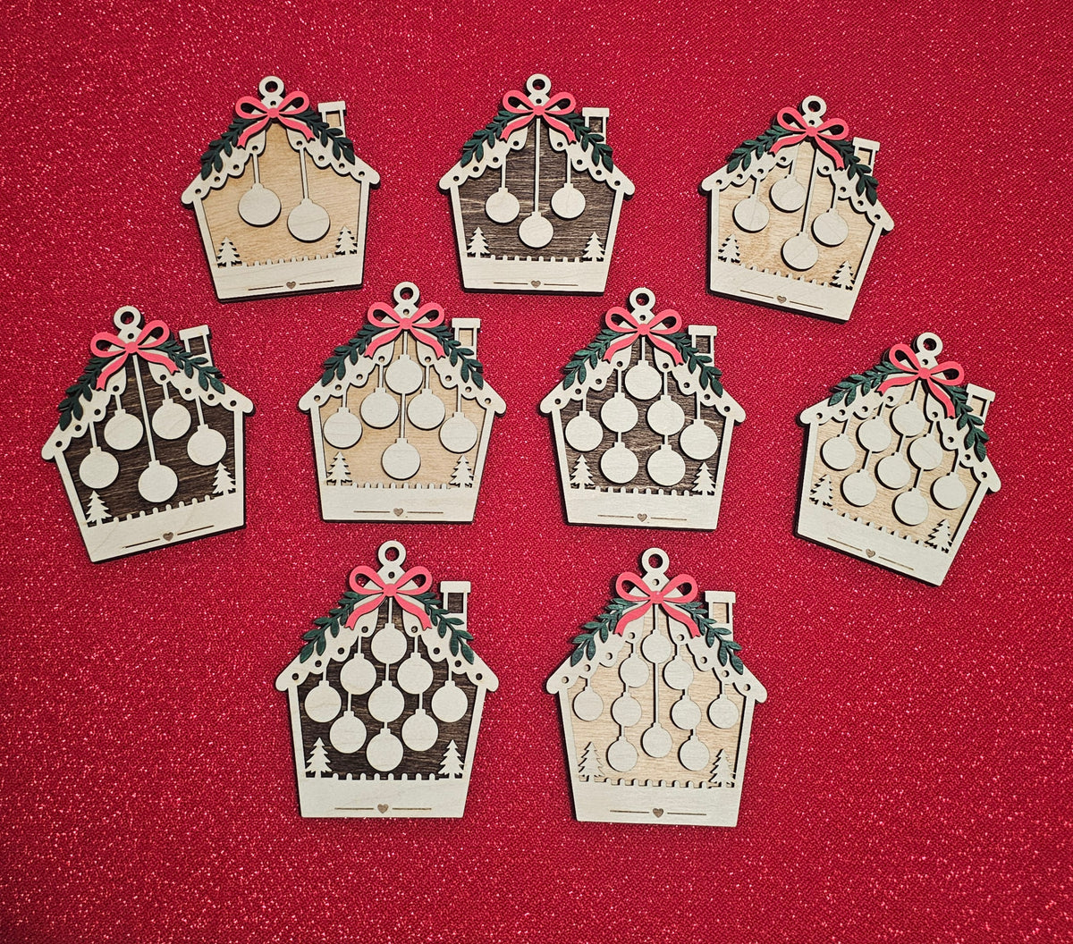 Seven laser-cut wooden Christmas ornaments shaped like houses with hanging ornaments, green holly accents, and red bows, arranged on a textured red glitter background.