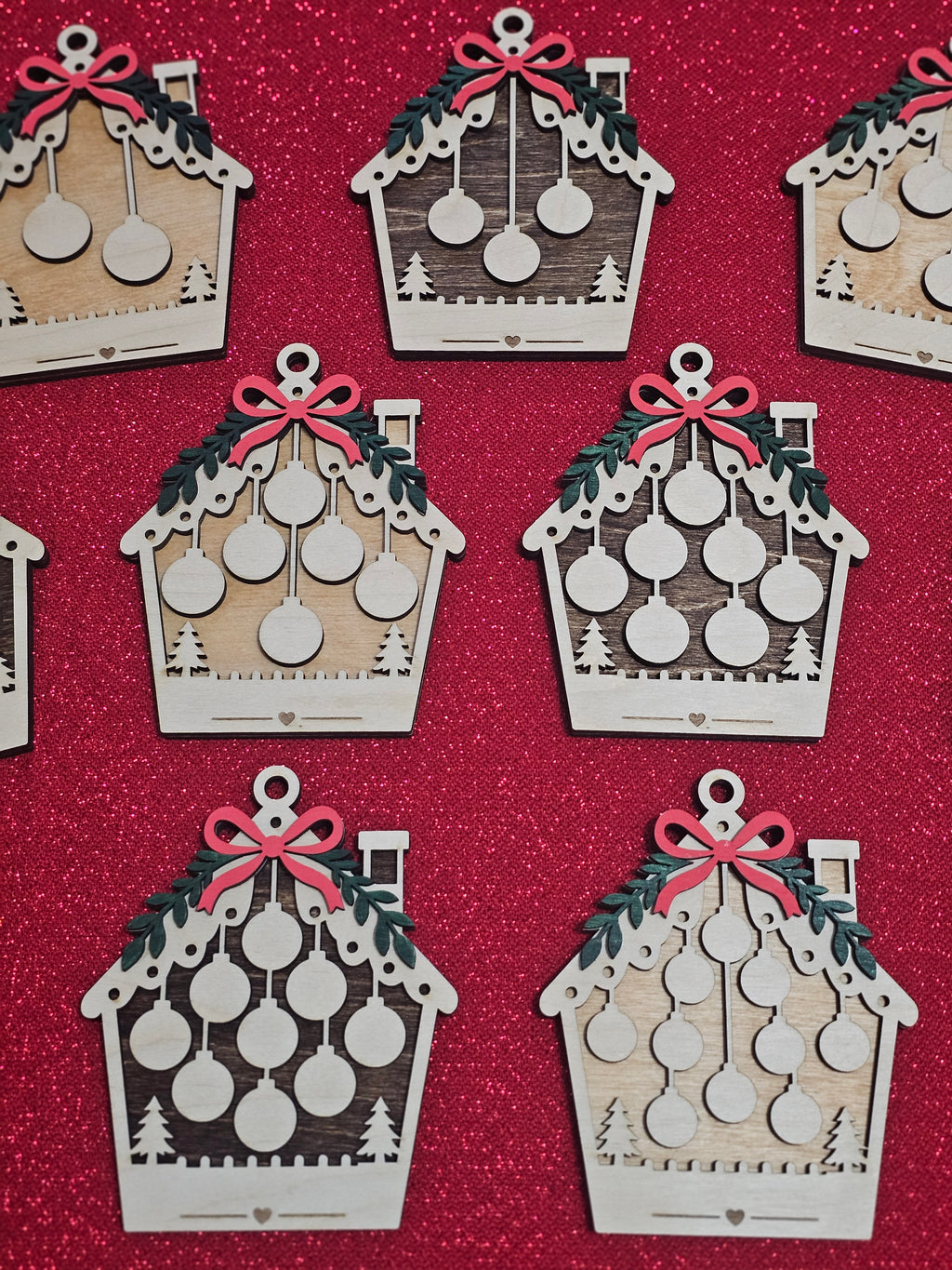 A set of wooden house-shaped Christmas ornaments with laser-cut details, featuring green holly leaves, red bows, and hanging silver ornaments, arranged on a textured red glitter background.