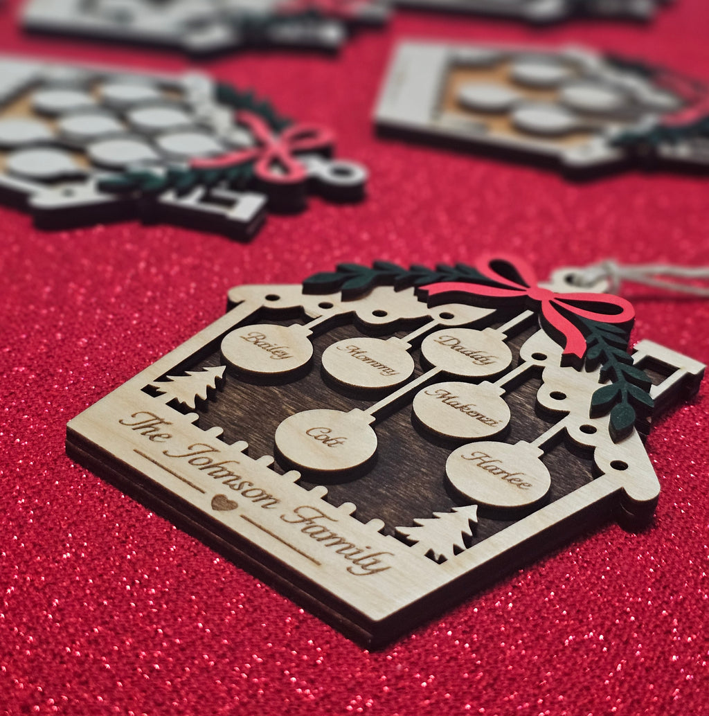 A personalized wooden house Christmas ornament engraved with 'The Johnson Family' and decorated with laser-cut baubles labeled with names, resting on a sparkling red glitter surface.