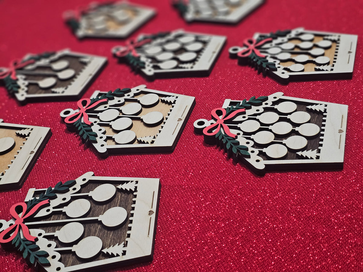 Laser-cut wooden Christmas ornaments with a house design, featuring circular cutouts, a red bow, green holly leaves, and pine trees, arranged on a textured red glitter background.