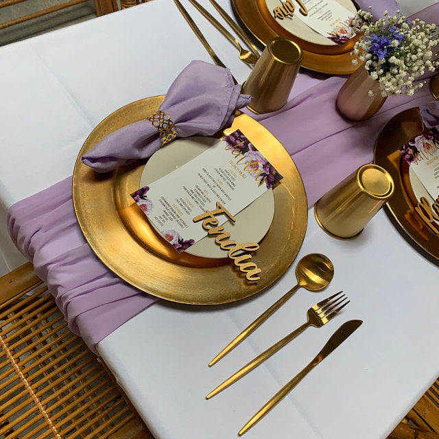 A styled table setting featuring gold charger plates, white dinner plates, a lavender napkin with a gold ring, gold flatware, and a lavender table runner on a white tablecloth.
